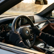 Driver's hands on BMW leather steering wheel with dashboard controls and carbon fiber accents in luxury car interior