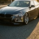 BMW sedan with lowered suspension and exposed chrome coilovers in wheel well cutaway, shot at low angle on asphalt in golden hour lighting.