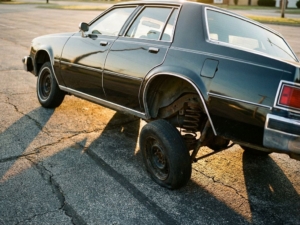 Car with severely compressed suspension leaning at extreme angle on asphalt, chassis nearly touching ground due to suspension failure.