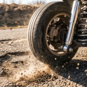 Car wheel and suspension system bouncing over pothole with tire airborne, chrome shock absorber visible, dust flying.