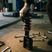 Mechanic's hand gripping aftermarket coilover suspension component with metallic spring coils and adjustment knobs in garage workshop