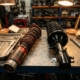 Worn coilover suspension unit and new factory strut assembly on mechanic's workbench with precision tools nearby