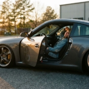 Lowered sports car with visible coilovers parked on asphalt, driver seated inside with open door in golden hour light.