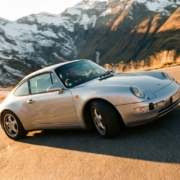 Silver Porsche 911 navigating mountain curve with driver visible through window, alpine landscape background, golden hour lighting.