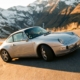 Silver Porsche 911 navigating mountain curve with driver visible through window, alpine landscape background, golden hour lighting.