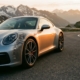Silver Porsche 911 cornering on mountain road with exposed suspension components and alpine scenery in background.