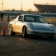 Silver Porsche 911 navigating orange traffic cones on asphalt during golden hour, captured mid-turn with dynamic body lean.