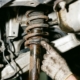 Mechanic pointing to worn car shock absorber with rust, oil leaks, and cracked rubber bushings in garage lighting.