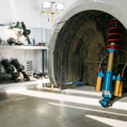 Close-up of aftermarket coilover suspension system with bright aluminum components in car wheel well, garage workbench visible.