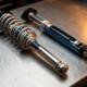 Two automotive struts on metallic workbench showing MacPherson and inverted designs with chrome and anodized finishes under dramatic workshop lighting