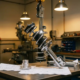 Chrome MacPherson strut assembly suspended upside-down in precision workshop, coil spring and damper catching golden light on steel workbench