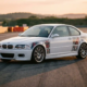 White BMW E46 M3 on race track with transparent cutaway showing red Intrax coilovers and suspension components in golden hour lighting