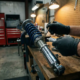Mechanic applying lubricant to chrome coilover suspension adjustment mechanism on garage workbench with tools in background