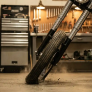 Motorcycle front fork compressing during hard braking with chrome suspension tubes telescoping inward, tire deformed against asphalt in garage workshop
