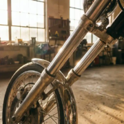 Motorcycle front fork assembly with angled wheel displaying caster geometry, chrome suspension components in garage workshop with golden lighting