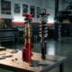 Red anodized motorsport coilover and black street coilover side by side on automotive workbench, showing internal damping mechanisms and spring configurations