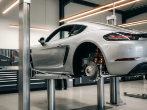 Silver Porsche 718 Cayman on hydraulic lift with rear wheel removed, exposing orange and black Intrax suspension components in modern garage