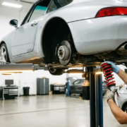 Silver Porsche 911 on hydraulic lift with mechanic inspecting red coilover suspension components in white garage workshop