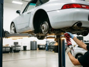 Silver Porsche 911 on hydraulic lift with mechanic inspecting red coilover suspension components in white garage workshop