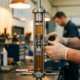 Engineer's hands adjusting a precision shock absorber mid-compression in automotive workshop, showing detailed internal damping mechanism and hydraulic chambers with chrome surfaces under warm lighting.