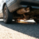 Car undercarriage scraping asphalt with visible suspension components, exhaust pipe, and orange sparks flying from metal contact