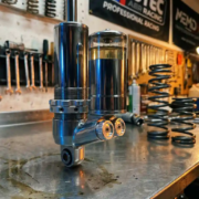 Chrome shock absorber with internal reservoir chamber on garage workbench surrounded by automotive tools and suspension parts