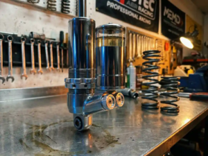 Chrome shock absorber with internal reservoir chamber on garage workbench surrounded by automotive tools and suspension parts