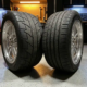 Two high-performance tires on polished aluminum wheels displayed side by side on garage floor, racing tire with aggressive tread next to street tire with deeper grooves